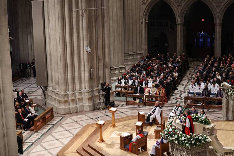 A imagem mostra o interior de uma catedral durante uma cerimônia. Há um altar ao centro， cercado por flores. Um grupo de pessoas está sentado em bancos， assistindo ao evento. À esquerda， há um grupo de pessoas em trajes formais， e à direita， um orador em vestes litúrgicas está falando. O ambiente é grandioso， com colunas altas e uma iluminação suave.