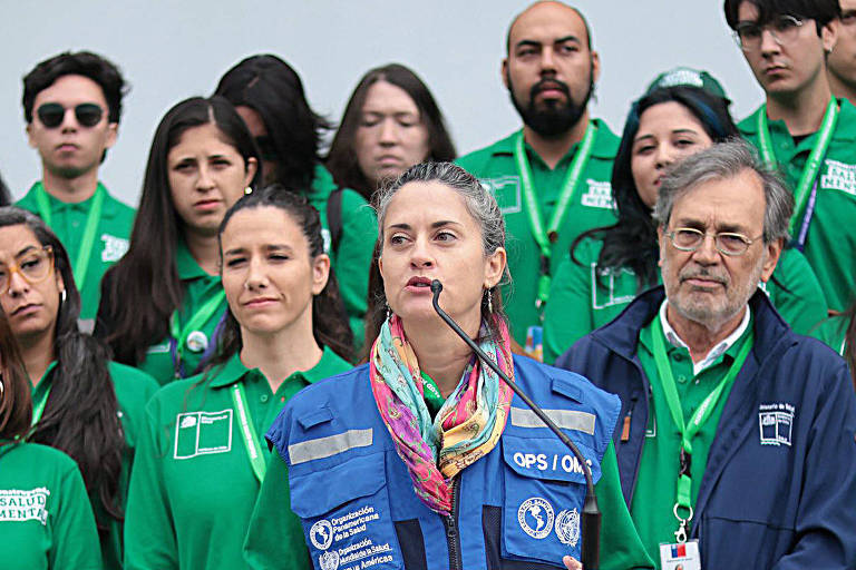 A imagem mostra um grupo de pessoas em uma coletiva de imprensa. No centro， uma mulher com cabelo liso e preso， vestindo uma jaqueta azul， está falando ao microfone. Atrás dela， há um grupo de pessoas， a maioria vestindo camisetas verdes， algumas com crachás visíveis. 