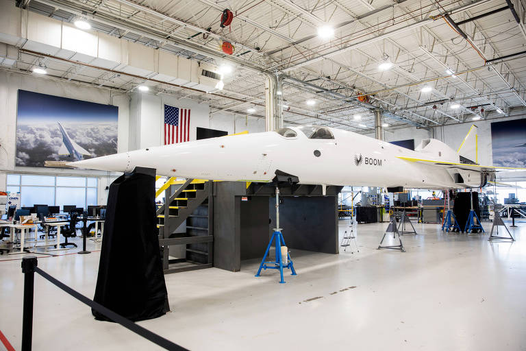 A imagem mostra um avião de fuselagem longa e estreita em um hangar. O avião está apoiado em suportes e coberto parcialmente por um pano preto. Ao fundo， há uma bandeira dos Estados Unidos e várias mesas de trabalho. O ambiente é bem iluminado， com um teto alto e paredes claras.
