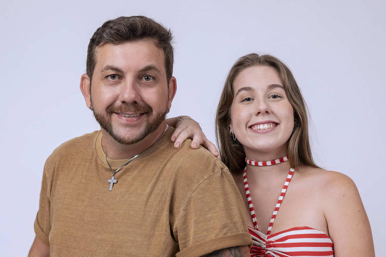 A imagem mostra um homem e uma mulher posando juntos. O homem está à esquerda, vestindo uma camiseta marrom e jeans, com um colar e um bracelete. Ele tem cabelo curto e barba. A mulher está à direita, usando um top listrado em vermelho e branco e uma saia longa combinando. Ela tem cabelo longo e solto, e está sorrindo. O fundo é branco.