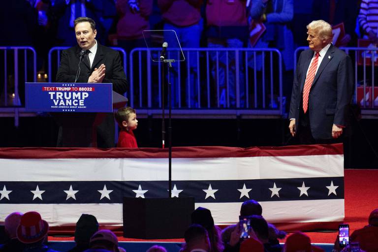 Imagem mostra o magnata Elon Musk falando em uma tribuna， enquanto o presidente dos Estados Unidos， Donald Trump， o observa， parado ao seu lado esquerdo. Musk fala para uma plateia， cujas cabeças e celulares apontados podem ser vistos na parte inferior da imagem. No púpito está escrito 039;Trump Vacance039;. 