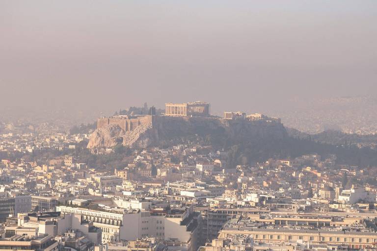 A imagem mostra uma vista panorâmica da cidade de Atenas, com a Acrópole visível ao fundo. A cidade é composta por muitos edifícios e casas, e a atmosfera parece estar um pouco enevoada, dificultando a visibilidade. A Acrópole, localizada em uma colina, é um dos principais pontos turísticos da cidade.