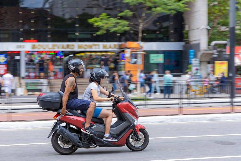 Uma motocicleta vermelha está em movimento na rua， com duas pessoas usando capacetes. O motorista está vestido com uma camiseta sem mangas e a passageira está usando um vestido. Ao fundo， há um edifício com várias pessoas e lojas visíveis.