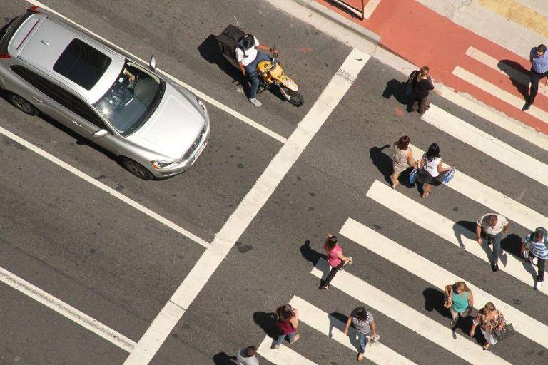 A imagem mostra uma vista aérea de um cruzamento de rua. Um carro prateado está parado na faixa de pedestres， enquanto várias pessoas estão atravessando a rua. Algumas pessoas estão caminhando em direção ao carro， enquanto outras estão na faixa de pedestres. O chão é marcado com faixas brancas e há uma área de calçada visível ao fundo.