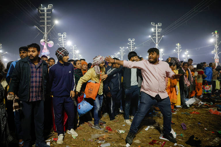 A imagem mostra uma multidão de pessoas em um evento noturno， com várias luzes ao fundo. Algumas pessoas estão vestindo roupas casuais， enquanto outras usam turbantes. Um homem em destaque， vestido com uma camisa rosa， parece estar gesticulando. O chão está coberto de lixo e há sacolas e objetos espalhados.