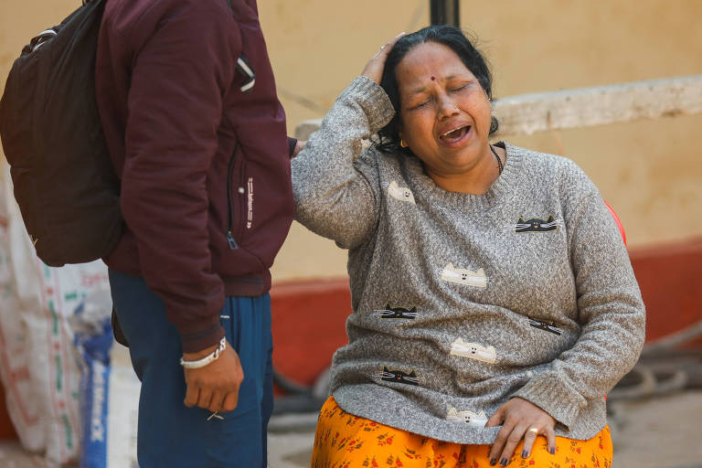 A imagem mostra uma mulher sentada， com uma expressão de dor emocional， segurando a cabeça com uma das mãos. Ela está vestindo um suéter cinza e uma saia laranja. Ao lado dela， um jovem está de pé， olhando para ela. O fundo é simples， com uma parede clara e alguns objetos ao redor.