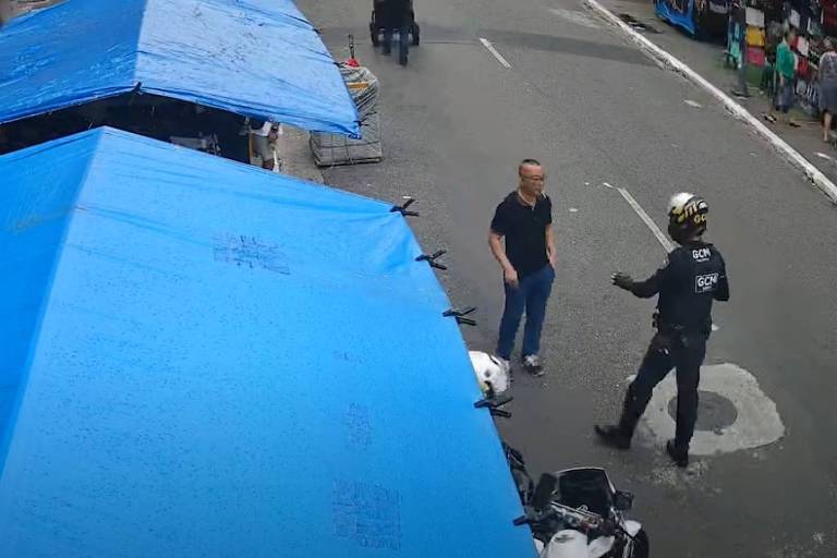 A imagem mostra uma rua com barracas azuis montadas. Um homem de camiseta preta está em pé conversando com um policial que usa um uniforme escuro e capacete. Ao fundo, há pessoas caminhando e um veículo estacionado. O ambiente parece ser urbano.