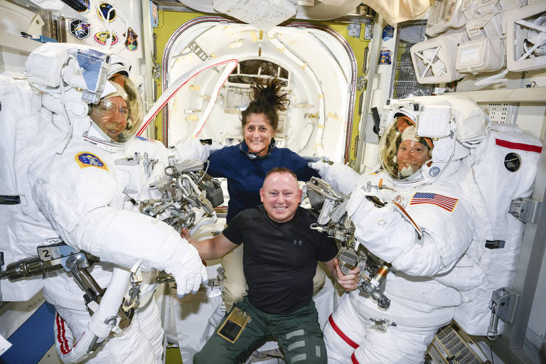 A imagem mostra quatro astronautas dentro da Estação Espacial Internacional. Dois deles estão vestidos com trajes espaciais brancos， enquanto um terceiro， no centro， está usando uma camiseta preta e calças verdes. A quarta astronauta， atrás do homem， está sorrindo e tem cabelo cacheado. O ambiente é interior da estação， com painéis e equipamentos visíveis ao fundo.