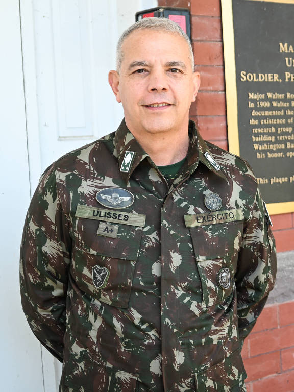 homem branco e com cabelo grisalho cortado rente à cabeça de uniforme militar com os braços cruzados atrás das costas