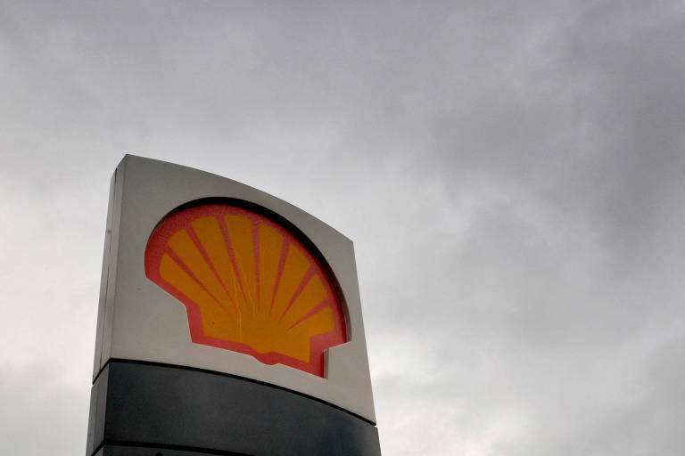 A imagem mostra um letreiro da Shell， com o logotipo característico em forma de concha， em cores amarelo e vermelho. O fundo é de um céu nublado， com nuvens cinzentas.