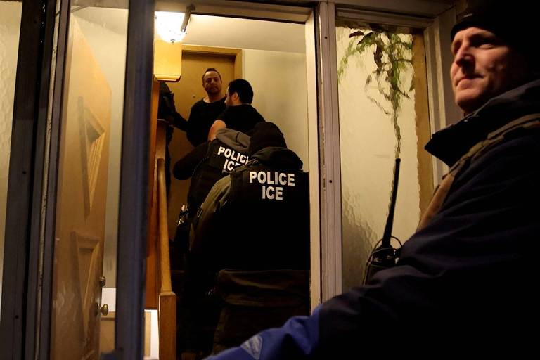 A imagem mostra uma cena de operação policial em uma residência. Há três agentes da polícia， todos vestidos com coletes que têm a inscrição 039;POLICE ICE039;. Um dos agentes está subindo as escadas， enquanto outro está em pé na porta， conversando com um homem que está dentro da casa. Um quarto agente está do lado de fora， observando a cena. A iluminação é baixa， sugerindo que a operação ocorre à noite.