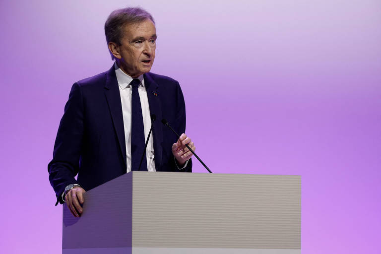 Imagem mostra Bernard Arnault， um homem em um terno escuro e que está falando em um microfone， posicionado atrás de um púlpito. O fundo é de uma cor roxa suave， criando um ambiente formal para a apresentação.