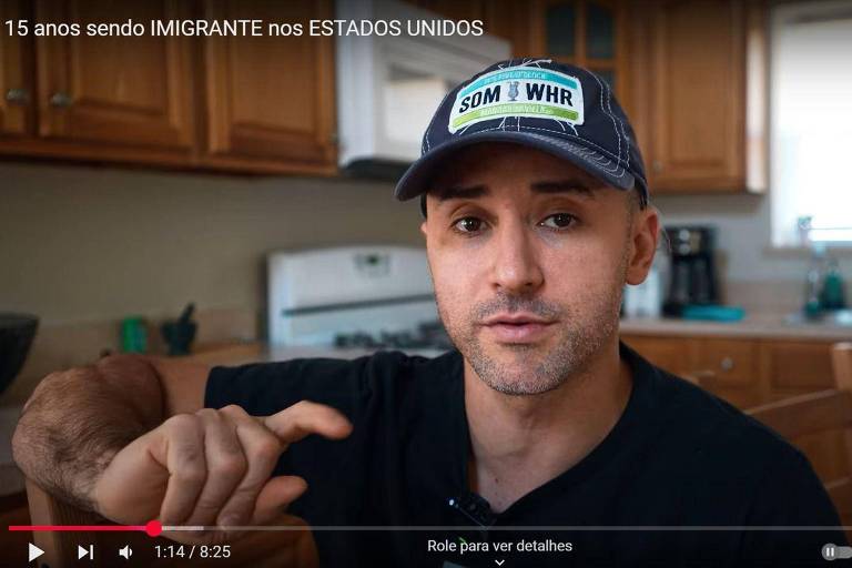 Um homem está sentado em uma cozinha， falando diretamente para a câmera. Ele usa uma camiseta preta e um boné azul com a inscrição 039;SOM amp; WHR039;. Ao fundo， há armários de madeira e uma pia com utensílios. O ambiente é bem iluminado.