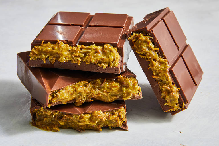 A imagem mostra várias barras de chocolate empilhadas. Cada barra tem uma camada de chocolate ao leite por fora e um recheio amarelo esverdeado， feito com pistache. Algumas barras estão parcialmente cortadas， revelando o recheio interno.