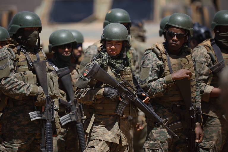 Um grupo de soldados em formação， todos usando uniformes camuflados e capacetes verdes. Eles estão armados e parecem estar em uma operação militar. O ambiente ao fundo é desértico， com veículos militares visíveis.