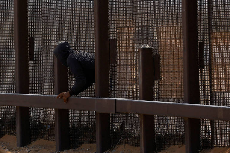 A imagem mostra uma pessoa vestindo um capuz， escalando uma cerca de metal. A cerca é alta e possui uma estrutura de malha， com um fundo de deserto visível. A pessoa está posicionada em um dos postes da cerca， com uma mão segurando a parte superior.