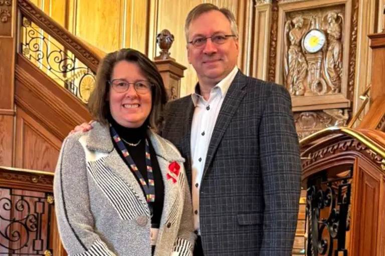 Duas pessoas estão posando para a foto em um ambiente interno decorado com madeira e detalhes ornamentais. A mulher à esquerda usa um blazer claro com listras e uma blusa preta, enquanto o homem à direita veste uma camisa branca e um blazer xadrez. Ambos estão sorrindo e parecem estar em um evento formal.