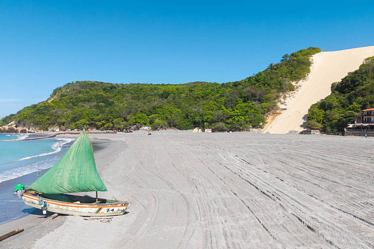 barco a vela， com vela de cor verde， atracado em faixa de areia com vegetação e corredor de areia ao fundo