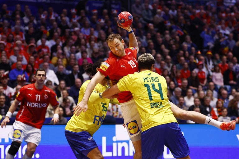 A imagem mostra um momento de um jogo de handebol， onde um jogador da equipe com uniforme vermelho， identificado como número 19， está saltando e segurando a bola acima da cabeça. Dois jogadores da equipe com uniforme amarelo， um deles com o número 14 e o outro com o número 15， estão tentando impedir o avanço do jogador vermelho. Ao fundo， uma multidão de torcedores pode ser vista， com muitos usando roupas em tons de vermelho.
