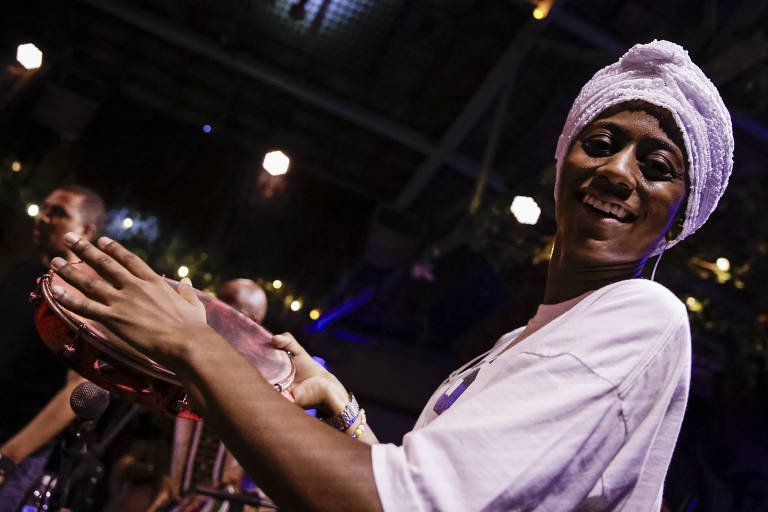 Uma mulher sorridente está tocando um pandeiro. Ela usa uma camiseta branca e um turbante branco na cabeça. Ao fundo, outros músicos estão visíveis, e a iluminação é suave, com luzes penduradas criando um ambiente festivo.