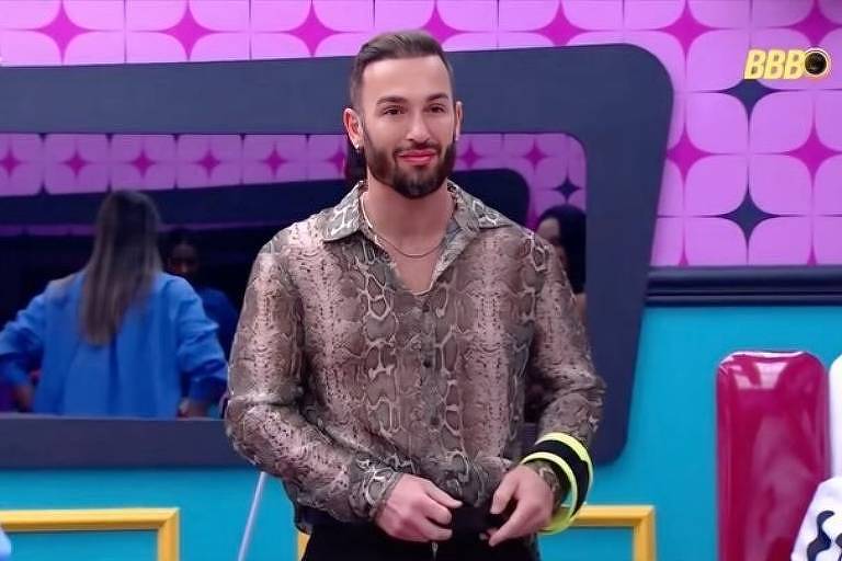 Um homem com cabelo liso e barba, vestindo uma camisa estampada em padrão de cobra, está em pé em um estúdio colorido. Ele sorri e parece estar se preparando para uma gravação. Ao fundo, há outras pessoas e um espelho refletindo a cena. O ambiente é decorado com paredes em tons de rosa e azul.