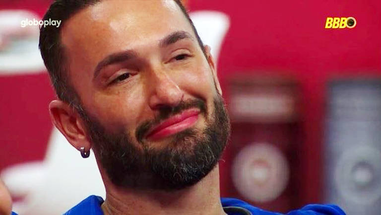 Um homem com barba e cabelo escuro, usando uma camiseta azul, está sorrindo levemente com uma expressão emocional. Ele parece estar em um ambiente interno, com um fundo vermelho e alguns objetos desfocados ao fundo. O homem tem um brinco na orelha esquerda e seus olhos estão levemente fechados, sugerindo uma sensação de felicidade ou nostalgia.