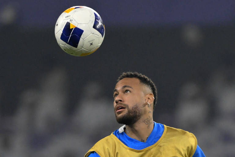 Um jogador de futebol está em um treino， olhando para uma bola que está flutuando no ar. Ele usa uma camiseta de treino azul e um colete amarelo. O fundo é desfocado， com pessoas e um ambiente de campo de futebol.
