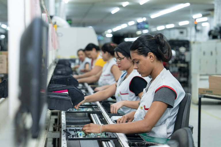 A imagem mostra várias mulheres trabalhando em uma linha de montagem em uma fábrica. Elas estão sentadas em cadeiras， concentradas em suas tarefas， que envolvem a montagem de componentes eletrônicos. O ambiente é bem iluminado e organizado， com mesas de trabalho e equipamentos visíveis ao fundo.