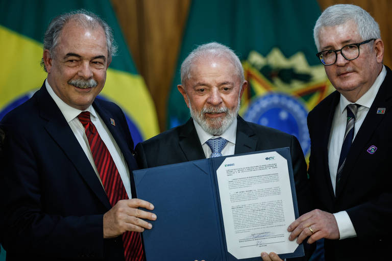 Três homens estão em uma cerimônia， segurando um documento. No meio está Luiz Inácio Lula da Silva， que está sorrindo e segurando o documento em destaque. Ao fundo， há bandeiras do Brasil. Os outros dois homens estão ao lado， um à esquerda e outro à direita， ambos sorrindo.