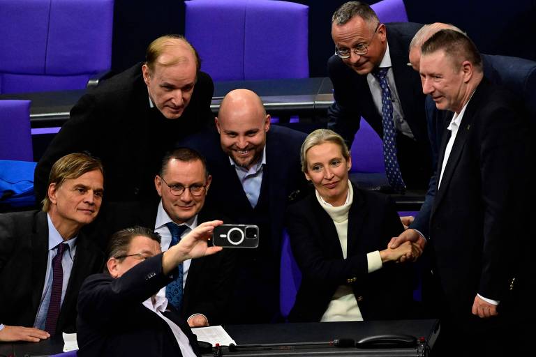 Um grupo de pessoas em um ambiente político， possivelmente em um parlamento， posando para uma selfie. Algumas pessoas estão sorrindo e olhando para a câmera， enquanto outras estão em segundo plano. O cenário é decorado com luzes e cores neutras， e há uma mesa na frente do grupo.