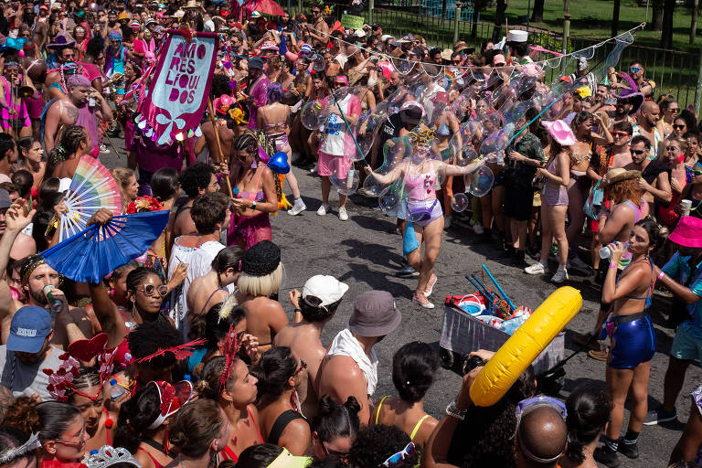 A imagem mostra uma grande multidão reunida em um evento festivo ao ar livre， com pessoas vestindo roupas coloridas e alegres. Algumas pessoas estão dançando e se divertindo， enquanto outras observam. O ambiente é vibrante， com muitos acessórios e bandeiras. O clima é de celebração e alegria.
