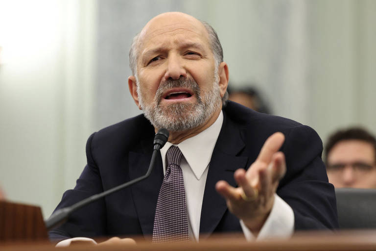 Imagem mostra Howard Lutnick， indicado para secretaria de Comércio dos EUA， um homem de cabelo grisalho e barba， vestido com um terno escuro e gravata， e que está falando em uma mesa durante uma audiência. Ele gesticula com a mão direita， parecendo expressar um ponto de vista ou responder a uma pergunta. Ao fundo， é possível ver outras pessoas sentadas， mas seus rostos não estão claramente visíveis.