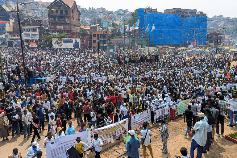 A imagem mostra uma grande multidão reunida em uma manifestação em uma área urbana. Muitas pessoas estão vestindo camisetas brancas e segurando faixas. Ao fundo， há edifícios e uma estrutura azul， possivelmente um andaime ou uma cobertura. O céu está claro e a cena parece ser durante o dia