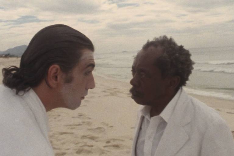 A imagem mostra dois homens em uma praia. O homem à esquerda tem cabelo escuro e liso, preso em um coque, e está usando uma roupa branca. Seu rosto está pintado de branco. O homem à direita tem cabelo crespo e grisalho, e está vestido com um terno branco. Eles estão se encarando intensamente, com o mar e o céu nublado ao fundo.