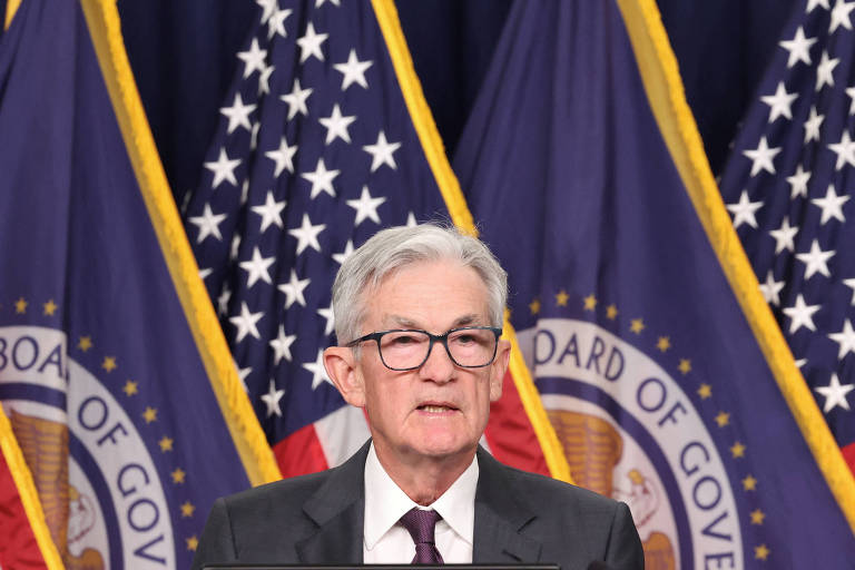 Um homem de óculos e cabelo grisalho está falando em um pódio， com bandeiras dos Estados Unidos ao fundo. As bandeiras têm o selo da Reserva Federal e estão dispostas em um padrão vertical.
