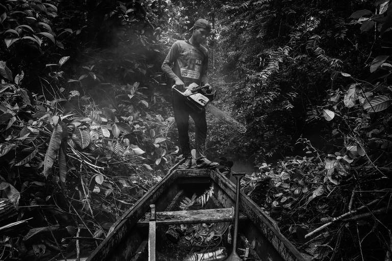 A imagem em preto e branco mostra um homem em pé na proa de uma canoa, cercado por uma densa vegetação de floresta. Ele segura um objeto nas mãos e parece estar em uma área isolada, com árvores e folhagens ao redor. A luz é suave, criando um ambiente misterioso.