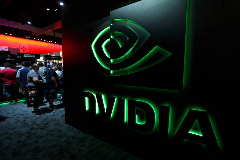 A imagem mostra um grande logotipo da NVIDIA em um fundo escuro. O logotipo é iluminado em verde e apresenta um design estilizado que lembra um olho. Ao fundo， é possível ver pessoas em um evento， com luzes vermelhas e outras cores iluminando o ambiente.
