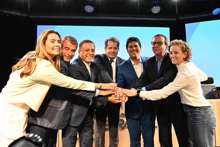 Um grupo de sete pessoas está em uma mesa， unindo as mãos em um gesto de colaboração. Eles estão sorrindo e vestindo roupas formais. Ao fundo， há um painel com a palavra 039;BRASIL039; e uma iluminação suave. A cena transmite um sentimento de união e trabalho em equipe.