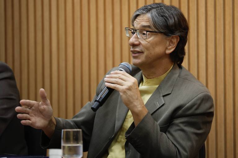 Um homem com cabelo liso e grisalho， usando óculos， está sentado em uma mesa. Ele segura um microfone com a mão direita e gesticula com a mão esquerda. O homem veste um paletó cinza e uma camiseta amarela. Ao lado dele， há um copo de água em uma mesa. O fundo é composto por painéis de madeira dispostos verticalmente.
