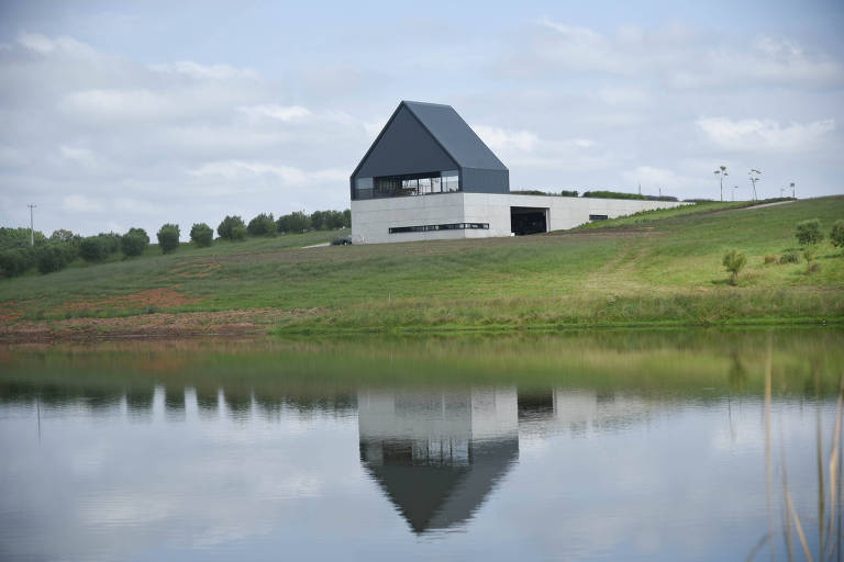 A imagem mostra uma casa moderna com telhado preto， localizada em uma colina verde ao lado de um lago. A casa possui uma estrutura retangular e janelas grandes， refletindo na água tranquila do lago. O céu está parcialmente nublado， e há uma linha de árvores ao fundo.