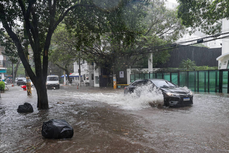 A imagem mostra uma rua inundada， com água cobrindo a calçada e parte da via. Um carro preto está passando pela água， levantando respingos. À esquerda， há um caminhão branco estacionado. Árvores estão visíveis ao longo da calçada， e há sacos de lixo flutuando na água.