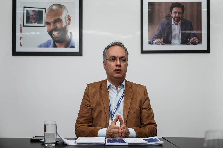 Padula， um homem de óculos e com cabelo grisalho， está sentado à mesa， gesticulando com as mãos em uma posição de oração. Ele usa uma camisa clara e um blazer marrom. Ao fundo， há duas fotos emolduradas de outros homens， Bruno Covas， prefeito de São Paulo que morreu em 2021， e Ricardo Nunes， vice de Covas e atual prefeito. Na mesa， há um copo de água e alguns documentos.