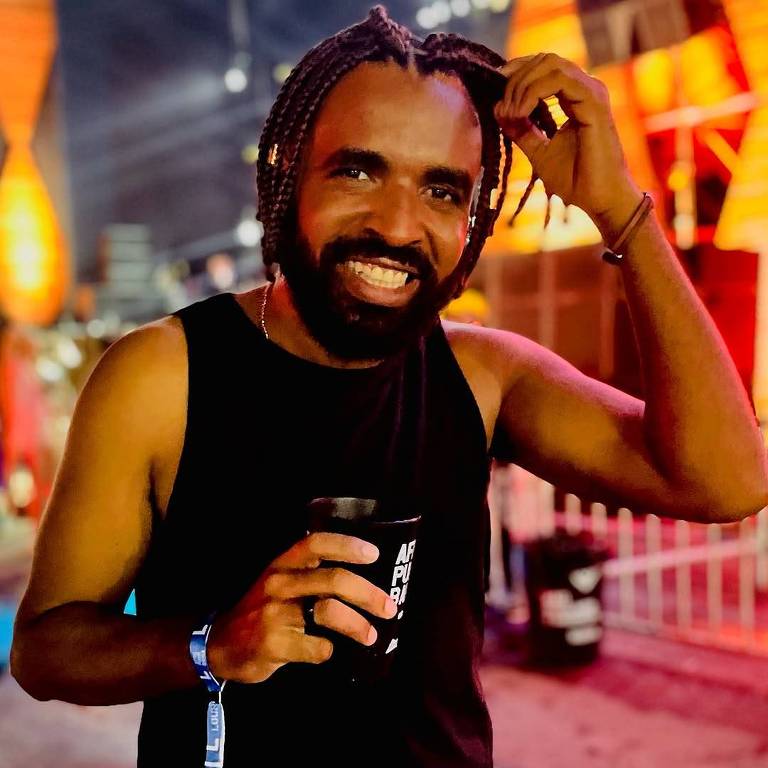 Um homem com cabelo trançado e barba， vestindo uma camiseta sem mangas preta， está sorrindo enquanto segura um copo. O fundo apresenta luzes coloridas e uma atmosfera de festa.