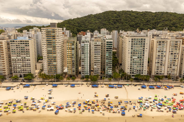 A imagem mostra uma vista aérea de uma praia com diversos guarda-sóis coloridos na areia. Ao fundo， há uma linha de edifícios altos e modernos， com uma montanha verde ao fundo. O céu está nublado， sugerindo um clima ameno.