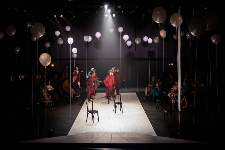 A imagem mostra um palco de teatro com várias pessoas em cena. No centro, há três cadeiras pretas dispostas em uma linha. Os atores estão vestidos com roupas coloridas, predominantemente em tons de vermelho e rosa, e estão se movendo em direção ao público. O fundo é escuro, com luzes focadas nos atores e balões brancos suspensos no ar. O público, visível nas laterais, observa a apresentação.