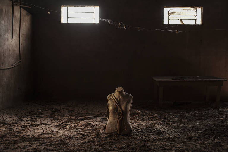 A imagem mostra um ambiente interno escuro e abandonado, com paredes e chão cobertos de sujeira. No centro, há um manequim sem braços, voltado para a câmera. Ao fundo, duas janelas pequenas permitem a entrada de luz, iluminando parcialmente o espaço. À direita, há uma mesa de madeira, também em um estado de abandono.