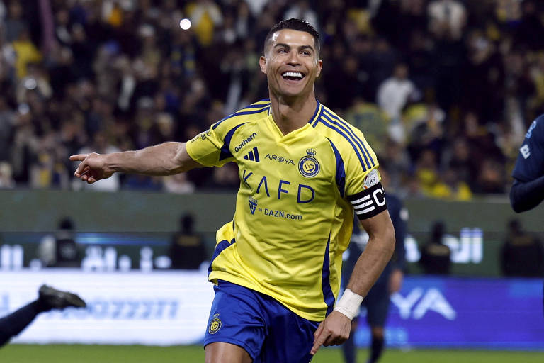 Um jogador de futebol está comemorando um gol. Ele veste uma camisa amarela com detalhes azuis e um símbolo de capitão no braço. O fundo mostra uma multidão de torcedores em um estádio.
