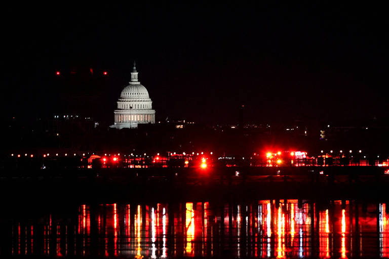 A imagem mostra o Capitólio dos Estados Unidos iluminado à noite， com um céu escuro ao fundo. Na parte inferior， há reflexos de luzes vermelhas na água， criando um efeito visual interessante