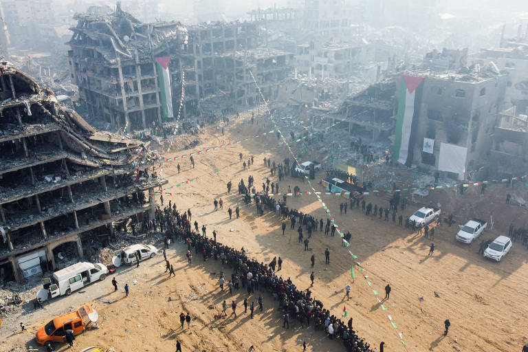 A imagem mostra uma área urbana devastada, com edifícios em ruínas e destroços visíveis. Há uma multidão de pessoas em fila, possivelmente esperando por ajuda ou suprimentos. Veículos de emergência estão presentes, e bandeiras com as cores da Palestina são visíveis em alguns edifícios. O ambiente parece empoeirado e nebuloso, sugerindo um cenário pós-conflito.