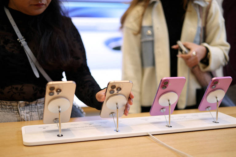 A imagem mostra uma exposição de smartphones em uma loja. Quatro celulares estão dispostos em uma base， com dois na cor rosa e dois em tons de bege. Uma pessoa à esquerda está segurando um celular， enquanto outra pessoa à direita está com as mãos próximas a um dos dispositivos. O fundo é desfocado， mas sugere um ambiente de loja.